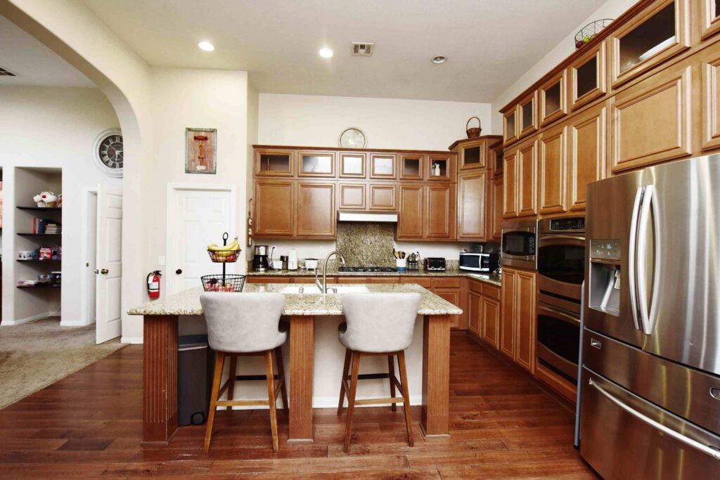 A modern kitchen interior featuring wooden cabinets, bar stools, and a central countertop with contemporary appliances.