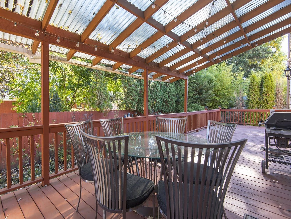 A cozy outdoor patio with a wooden pergola and glass table set amidst lush greenery.