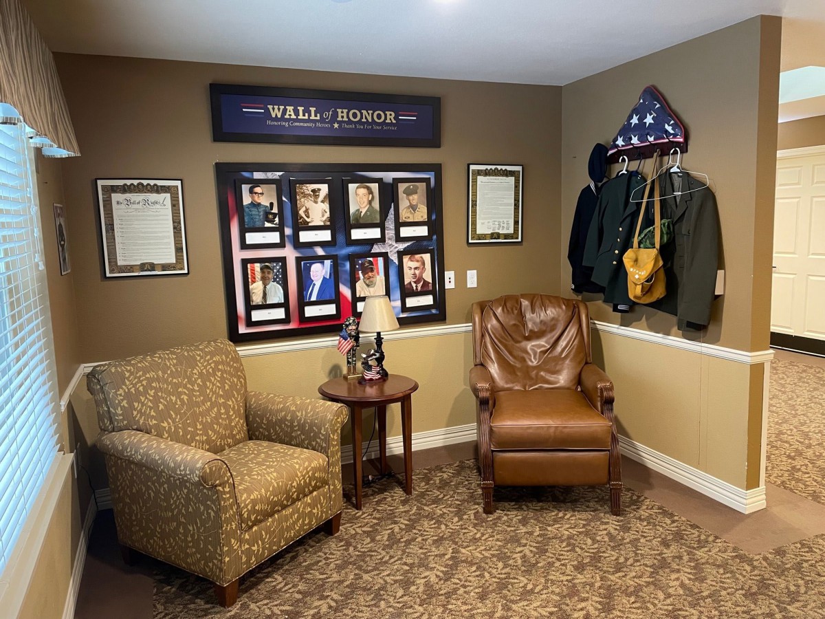 A cozy room featuring military memorabilia, including a framed 'Wall of Honor' display, uniforms, and a folded flag. Two comfortable chairs and a side table complete the setup.