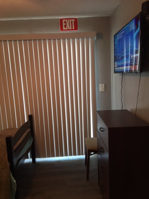 A small room with vertical blinds, a mounted TV, and furniture including a chair and dresser.