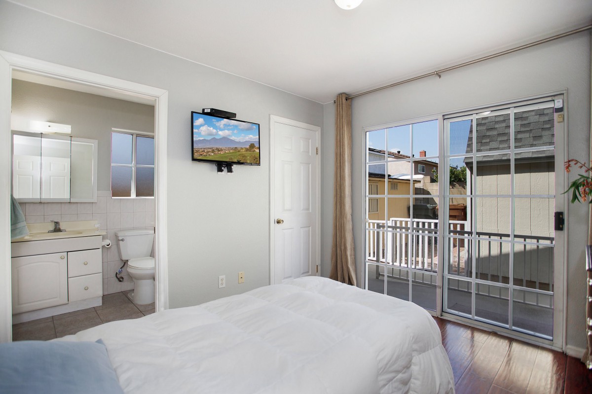 A modern, minimalist bedroom featuring a TV on the wall, sliding doors leading to a balcony, and a neatly made bed.