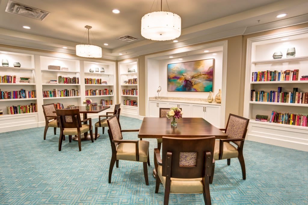 A well-lit library with bookshelves, tables, and chairs, featuring modern artwork on the wall.