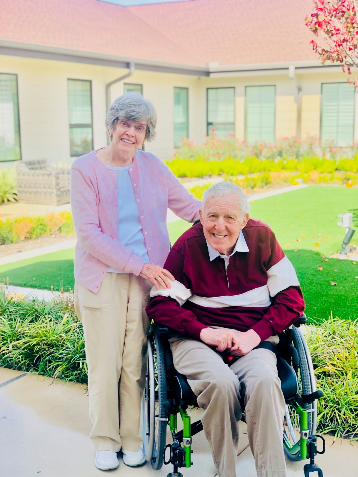 An elderly couple enjoying time together in a garden, with one seated in a wheelchair.