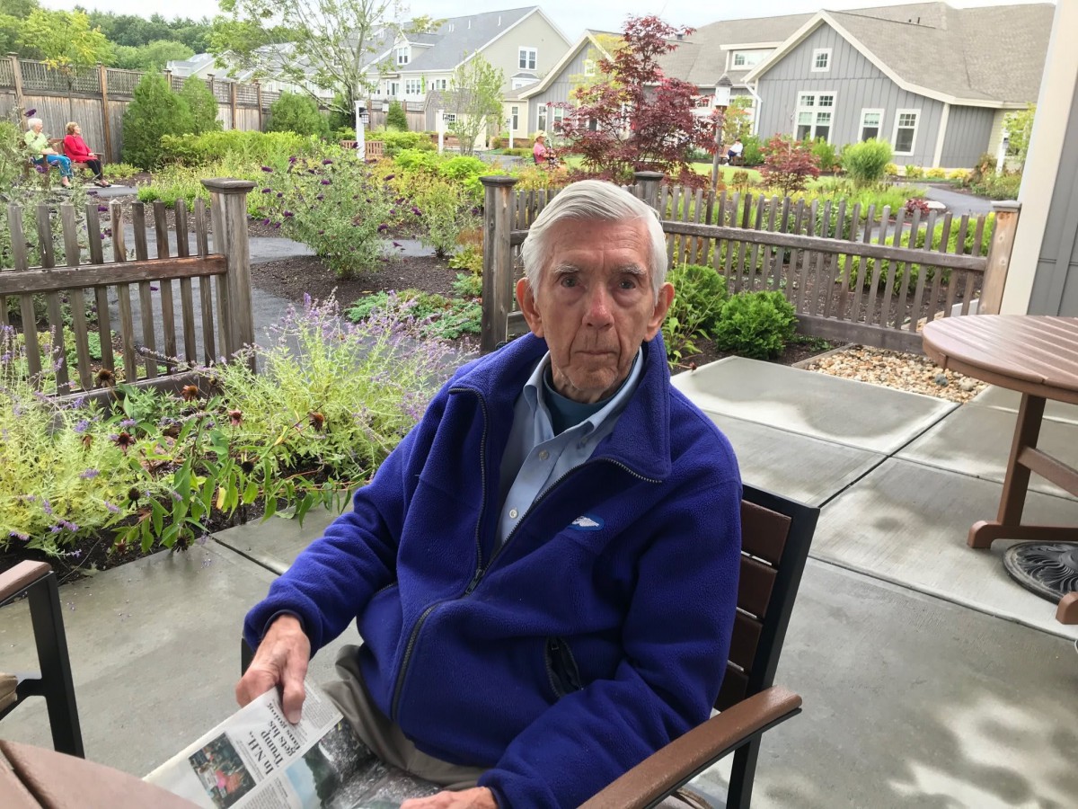 An elderly man sitting on a patio chair, holding a newspaper, with a garden in the background.