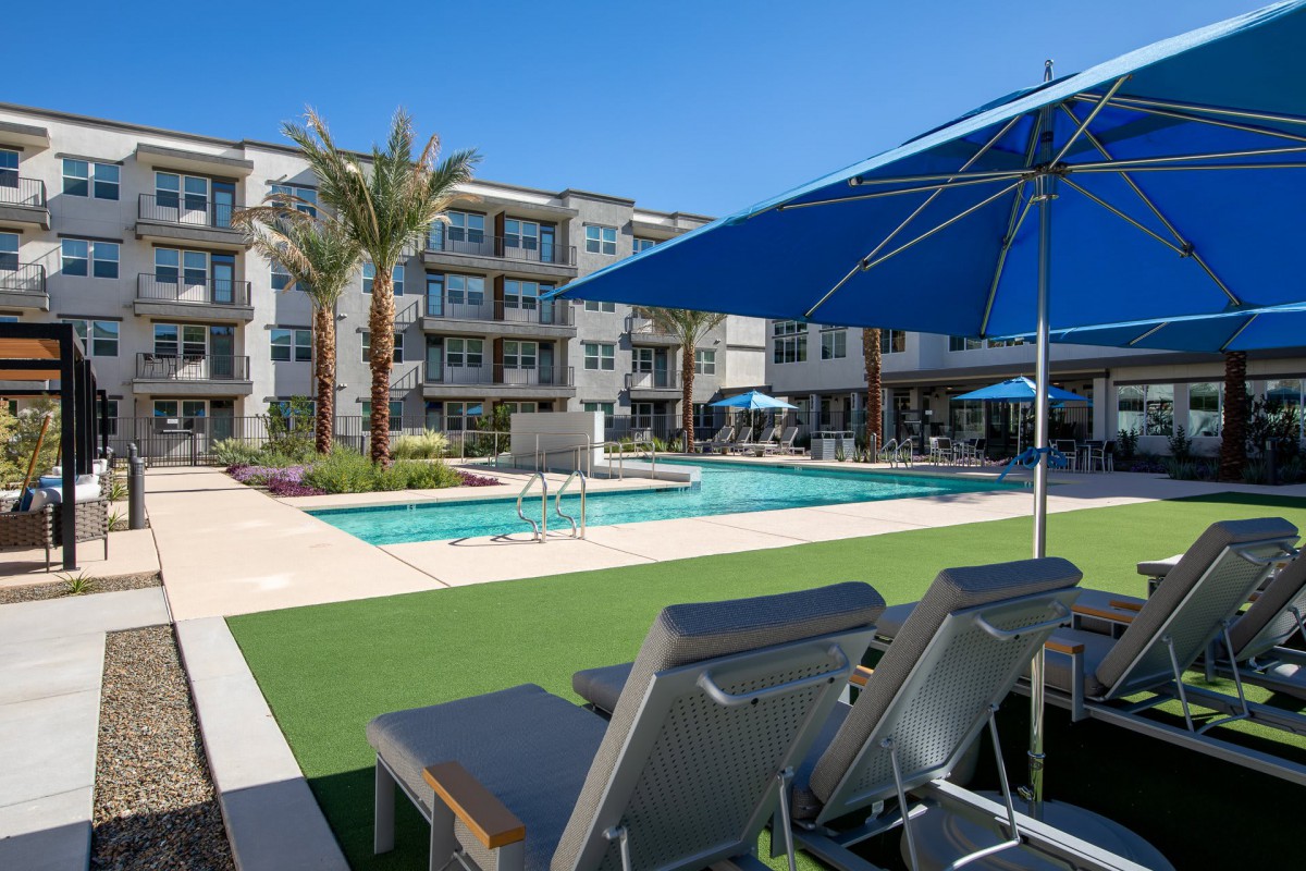 A modern apartment complex featuring a pool with surrounding lounge chairs and umbrellas on a sunny day.