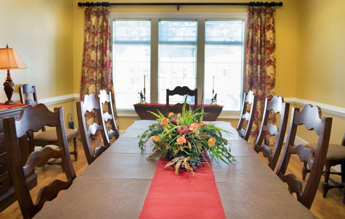 A well-decorated dining room featuring a long table with a floral centerpiece and matching chairs, framed by large windows with patterned curtains.