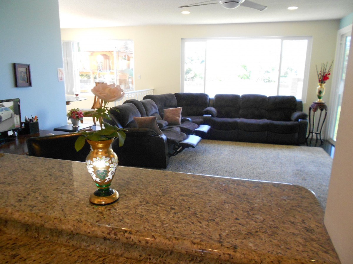 A cozy living room with a large sofa set, a vase of flowers on the counter, and sunlight streaming through the windows.