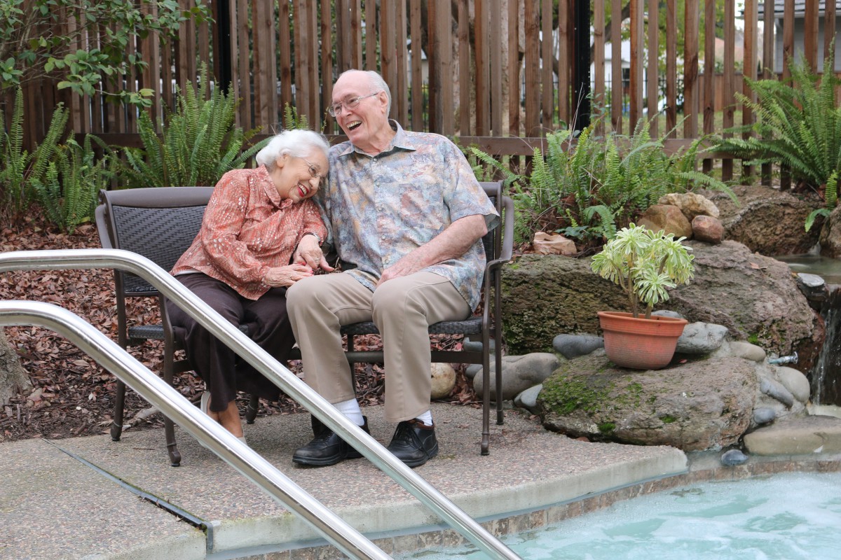 An elderly couple sits by the poolside, sharing a joyful moment outdoors.