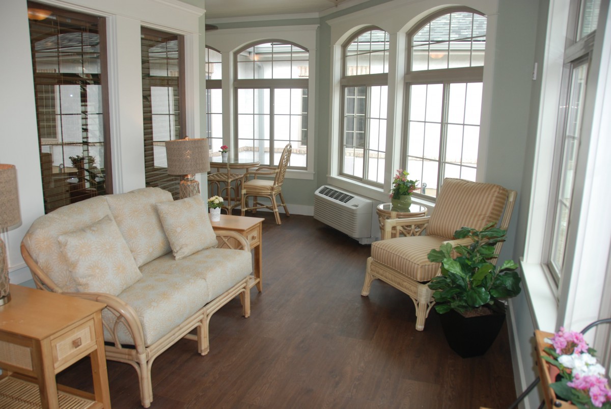 A cozy living room with ample natural light, featuring comfortable furniture, large windows, and indoor plants.