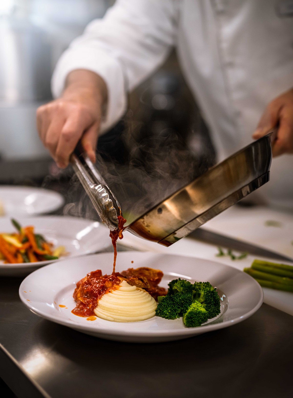 A chef skillfully plates a dish with sauce, served alongside mashed potatoes and broccoli.