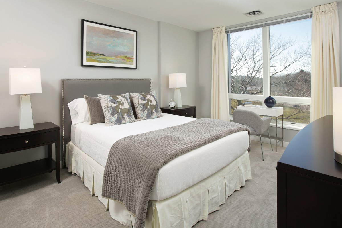 A modern bedroom featuring a neatly made bed with neutral-colored bedding and decorative pillows, complemented by a bedside lamp, window view, and a framed painting.