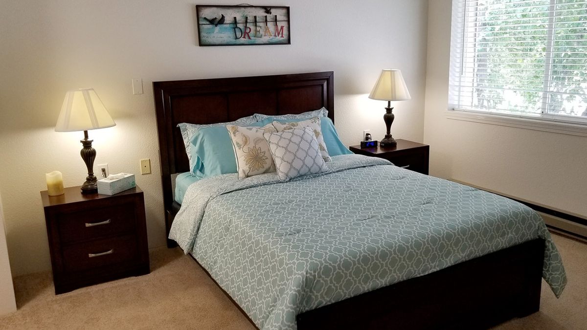 A cozy bedroom featuring a neatly made bed with blue bedding, decorative pillows, and a nightstand with a lamp.
