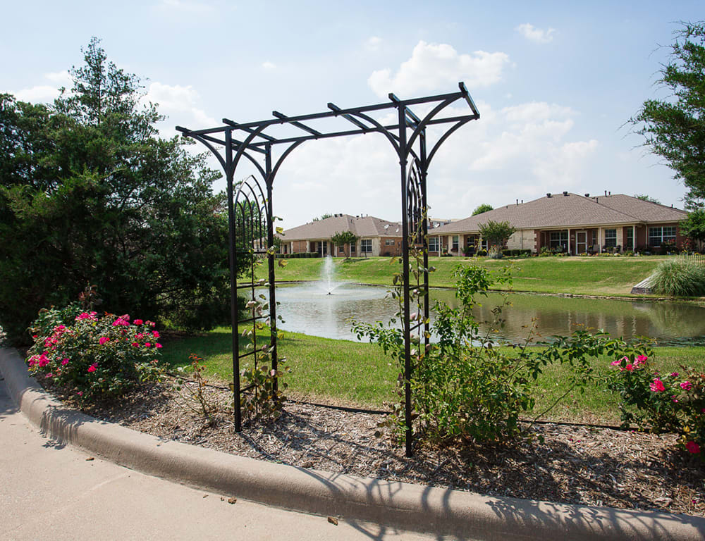 A decorative metal archway stands in a garden overlooking a pond with fountains and residential homes in the background.