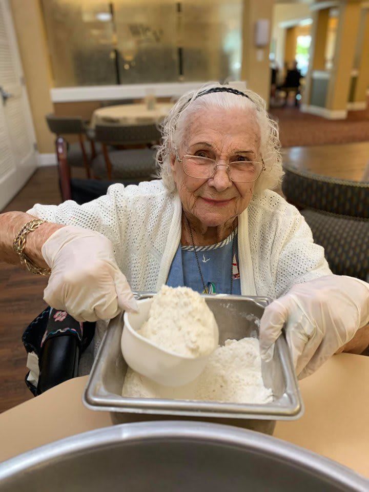 An elderly woman happily scooping flour in a kitchen setting.