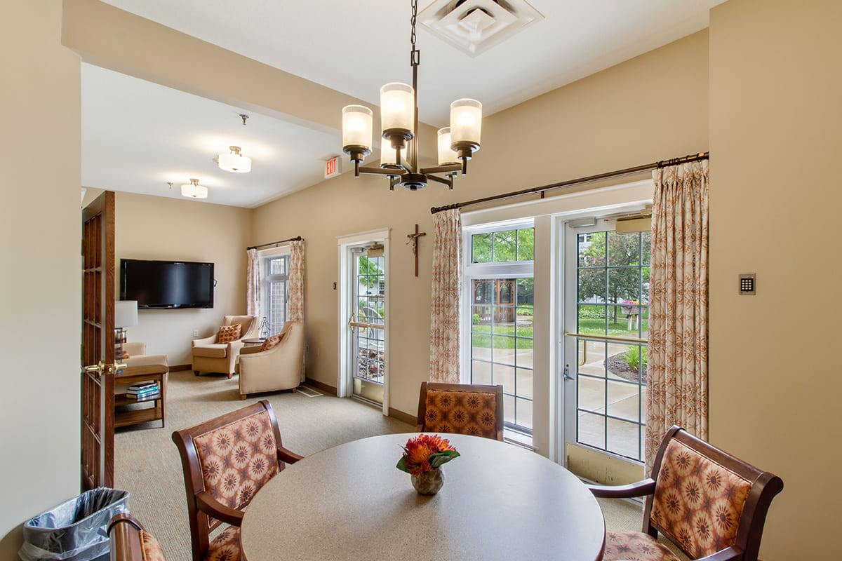 A cozy living room with a dining area, featuring a chandelier, comfortable chairs, and large windows overlooking a garden.