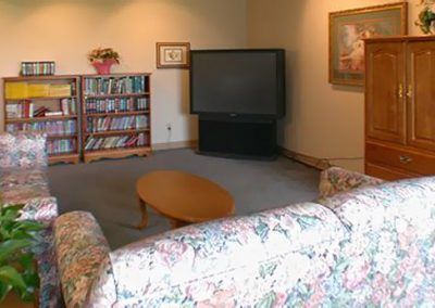 A cozy living room featuring a large television, a floral-patterned couch, a bookshelf filled with books, and a wooden coffee table on a carpeted floor.