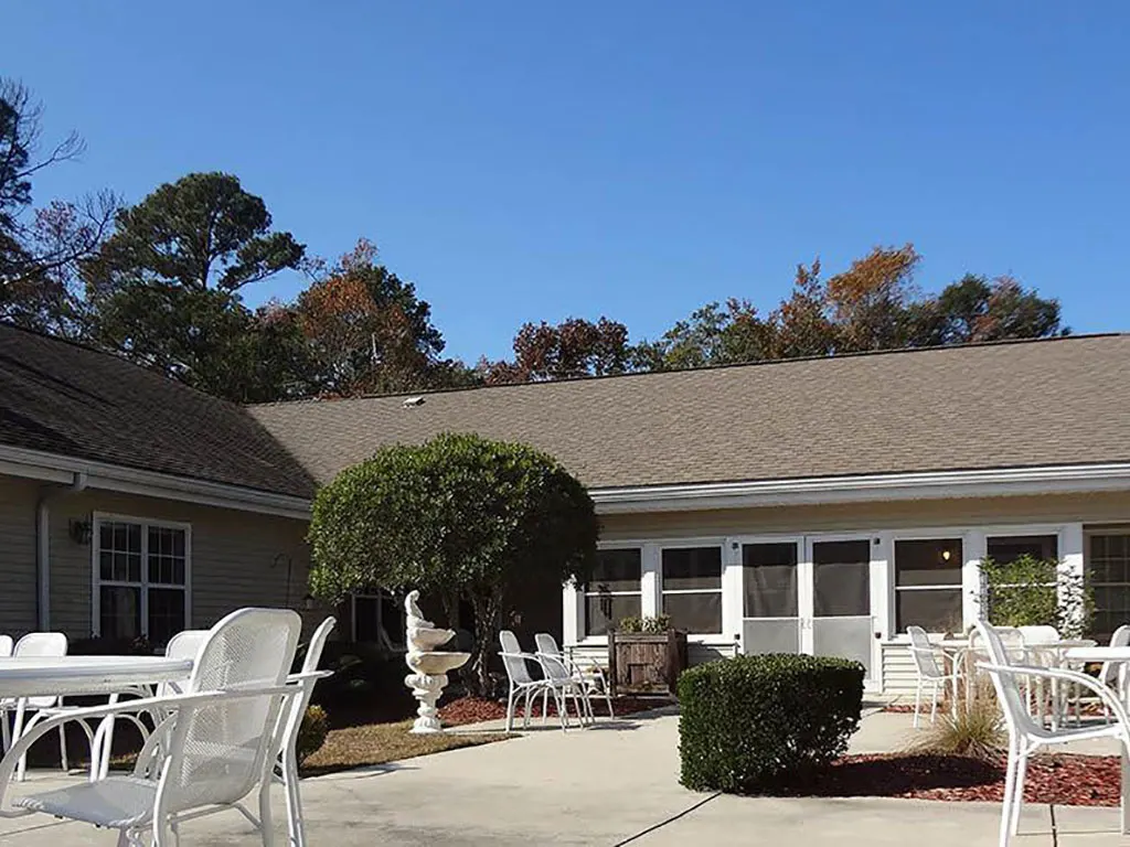 An outdoor patio area with tables and chairs, surrounded by bushes and trees under a clear blue sky.