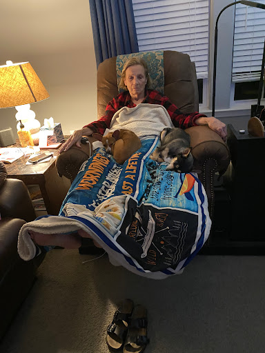 A person relaxing in a chair with three dogs snuggled under a colorful blanket.