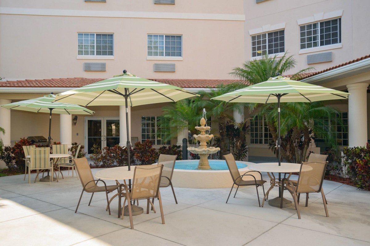 A patio area with tables and chairs shaded by umbrellas, set near a fountain and surrounded by palm trees.