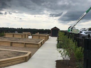 A rooftop garden with wooden planters under a cloudy sky, featuring a clear pathway and construction equipment in the background.