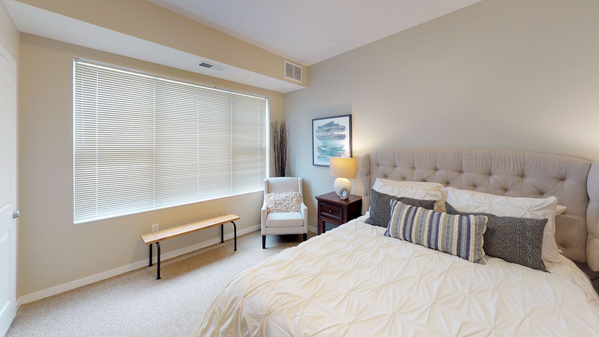 A cozy and well-lit bedroom with neutral colors, featuring a large window, a comfortable bed, an armchair, and tasteful decor.