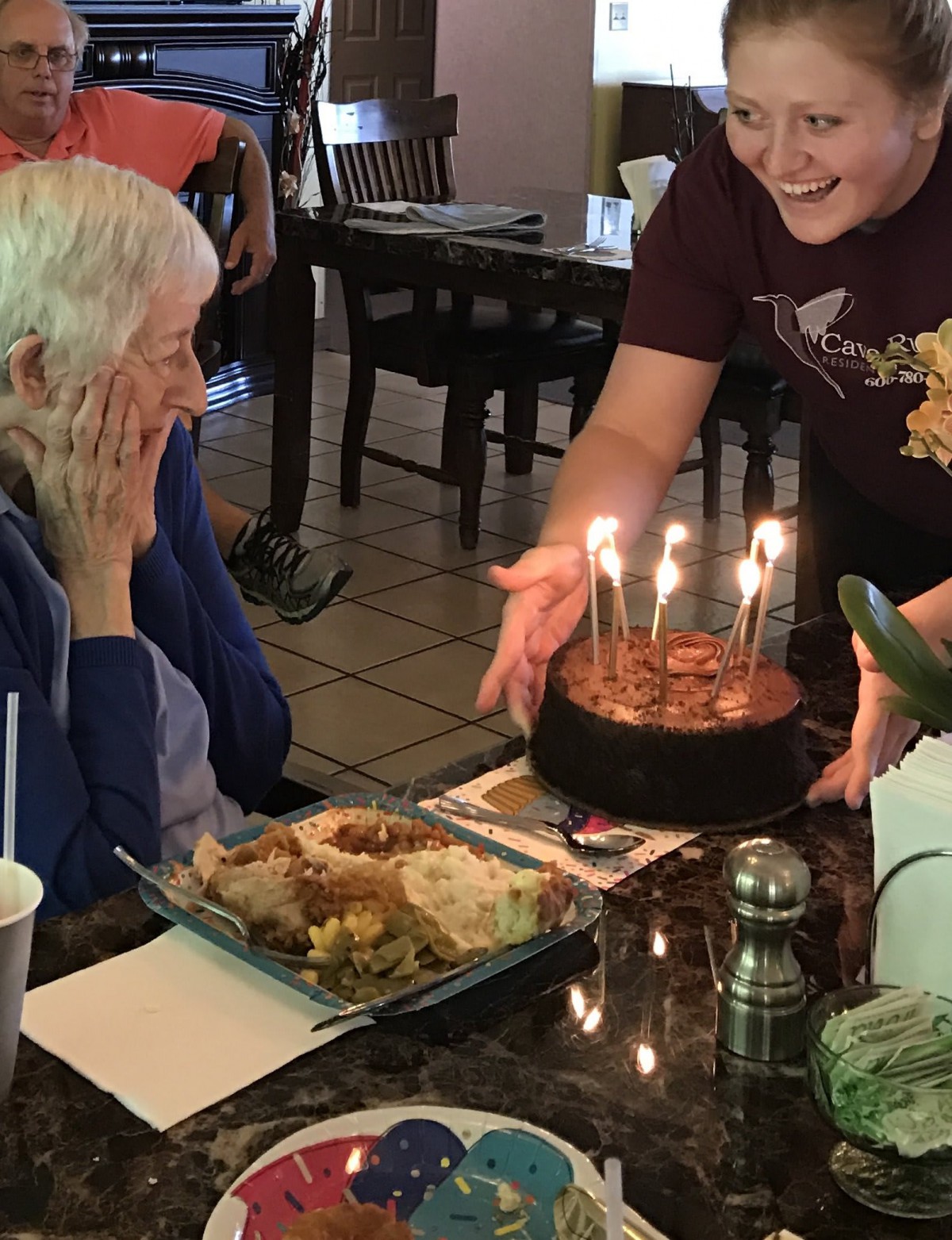 An elderly person smiling as a birthday cake with lit candles is presented in a restaurant setting.