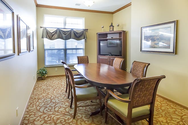 A small conference room with an oval wooden table surrounded by chairs, featuring a television, window with drapes, and patterned carpet.