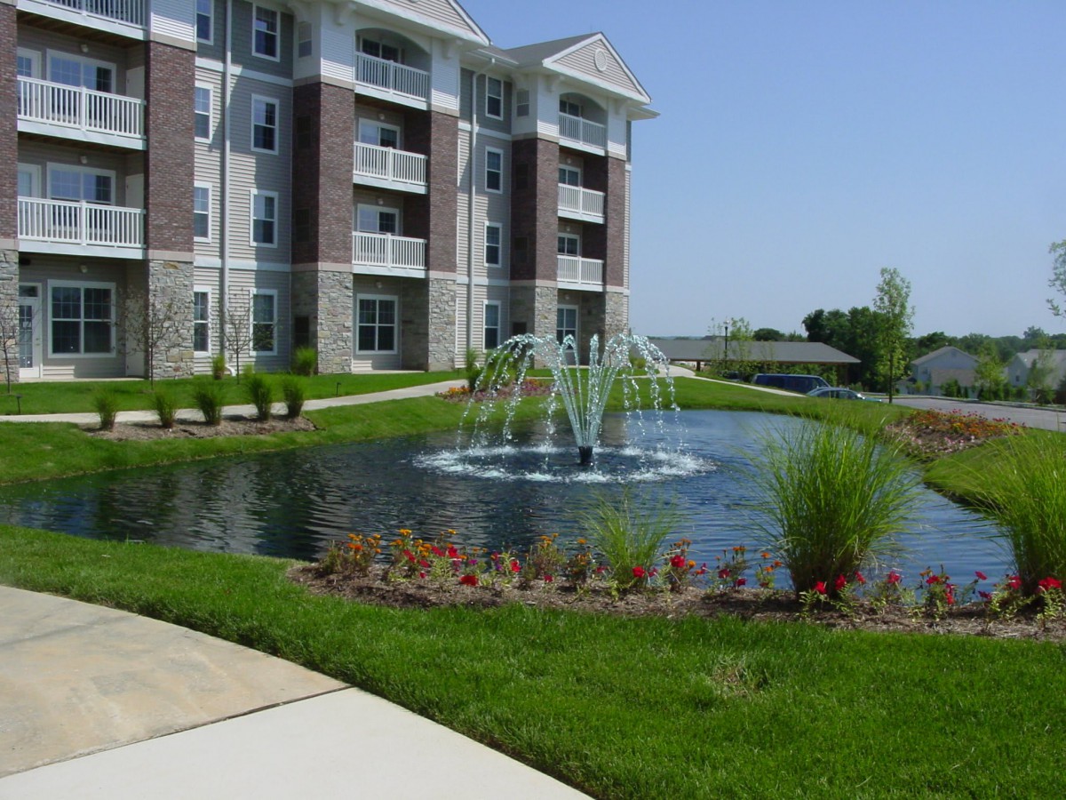 A modern apartment building with a landscaped pond and fountain in the foreground.