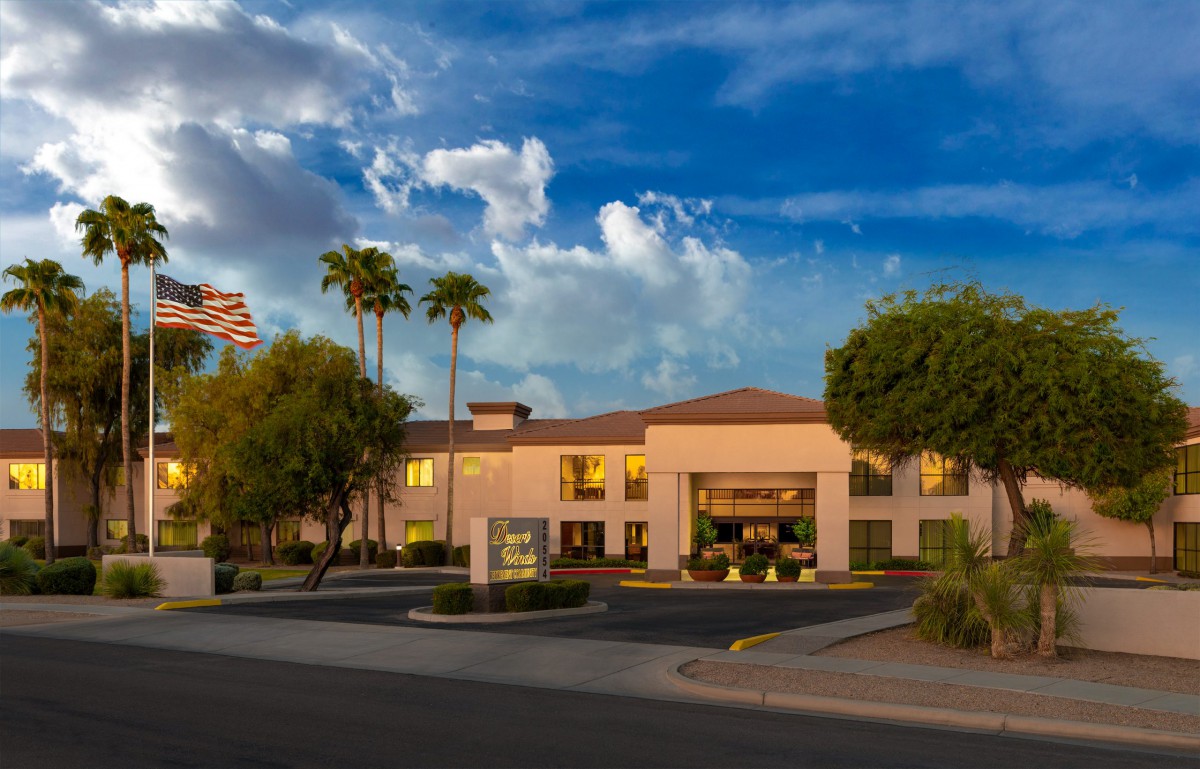 A hotel building surrounded by palm trees, with a parking lot in the foreground under a sunset sky.