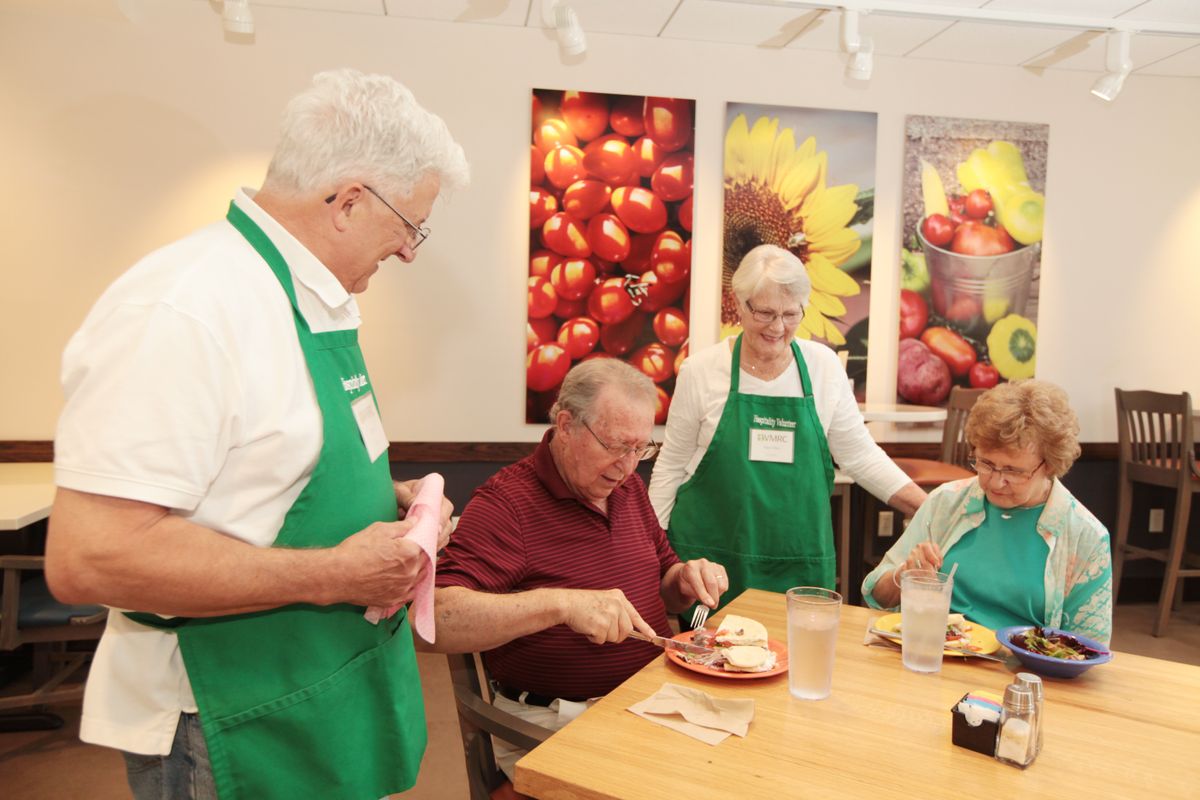 Volunteers wearing green aprons serve and socialize with seniors at a community event.