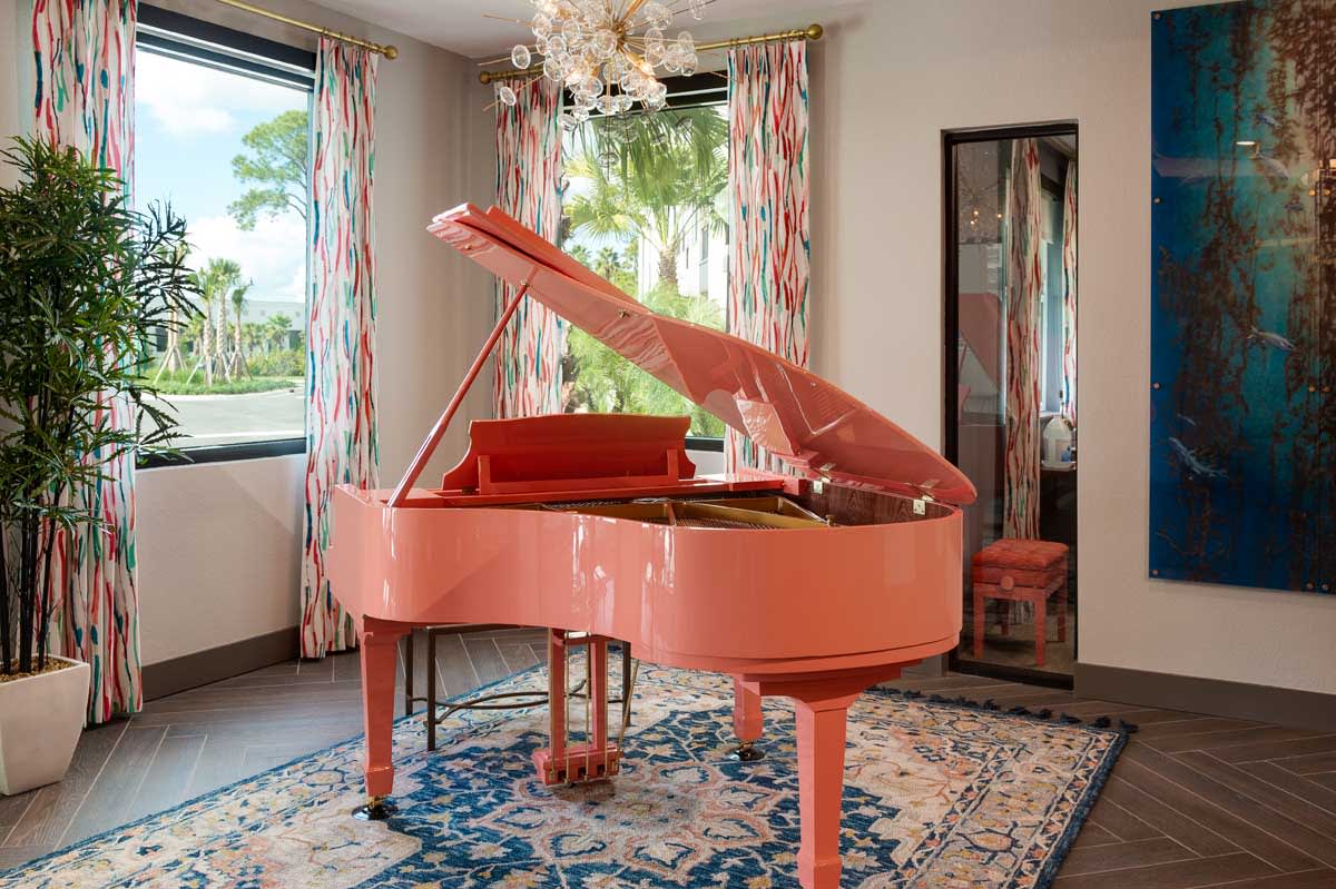 A stylish living room features a pink grand piano with a chandelier overhead and patterned curtains framing the windows.