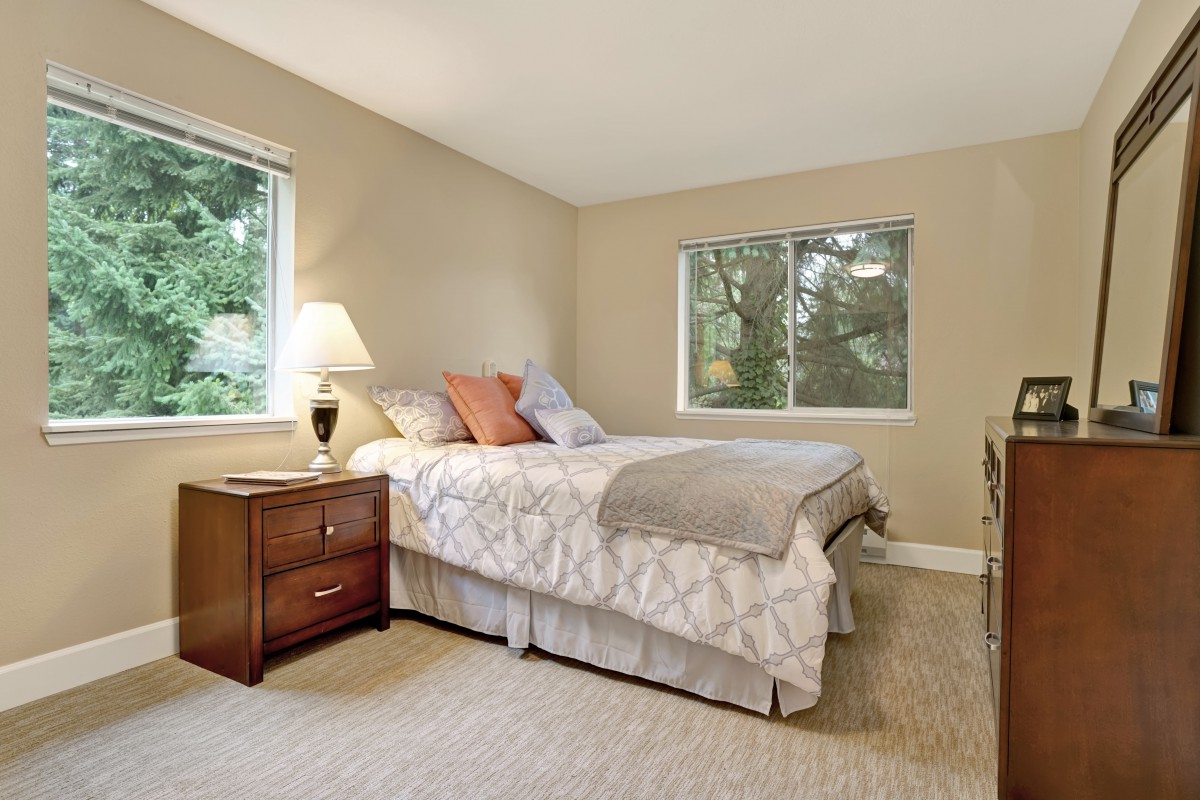 A cozy bedroom with a neatly made bed, two windows, and a nightstand with a lamp.