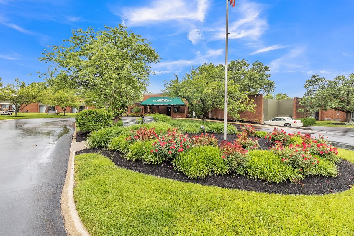 A beautifully landscaped area with a flagpole and a building in the background, surrounded by lush greenery and colorful flowers under a bright blue sky.