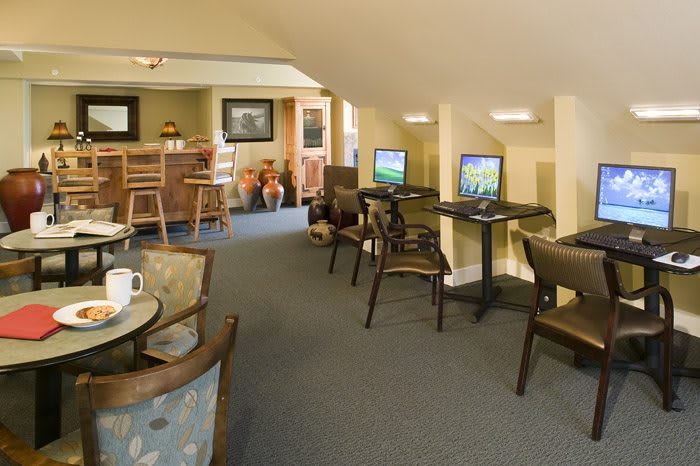 A comfortable lounge area featuring computers and a small bar section, with various chairs and tables arranged for guest use.