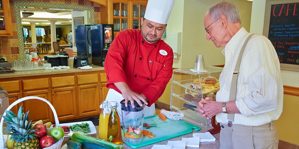 A chef in a red uniform demonstrates how to use a blender with fresh ingredients to an interested audience member in a kitchen setting.