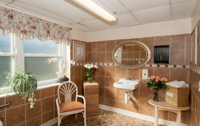 A cozy bathroom interior featuring brown tiles, a round mirror, a chair, and decorative flowers.