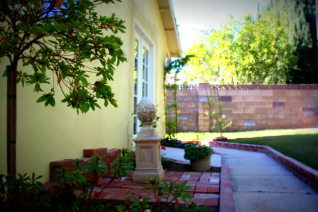 A scenic garden view featuring a pathway beside a yellow house with lush greenery and a decorative statue.