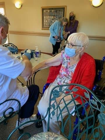 An elderly woman wearing a mask sits in a wheelchair and interacts with another person indoors.