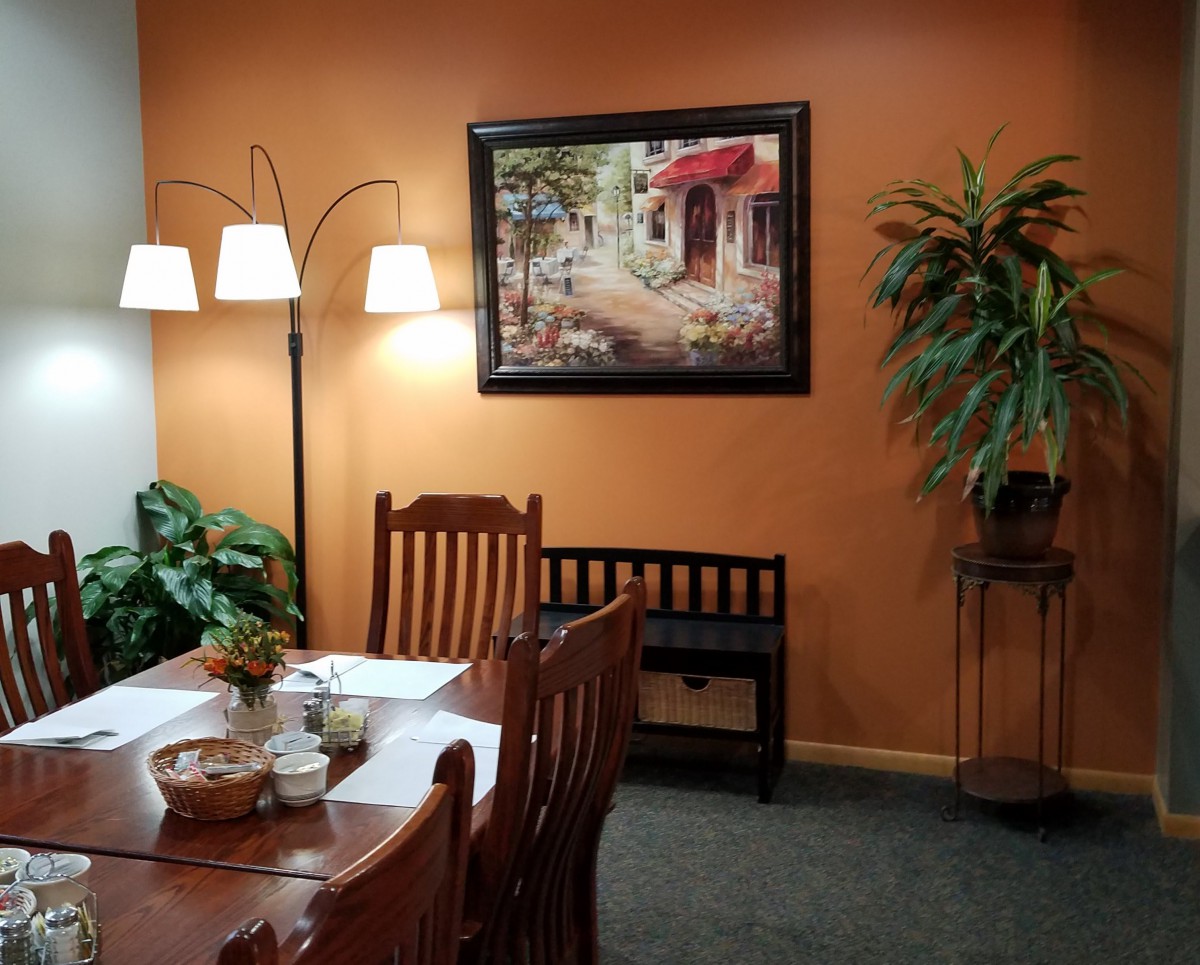 A cozy dining area with a wooden table and chairs, adorned with plants and a painting on an orange wall.
