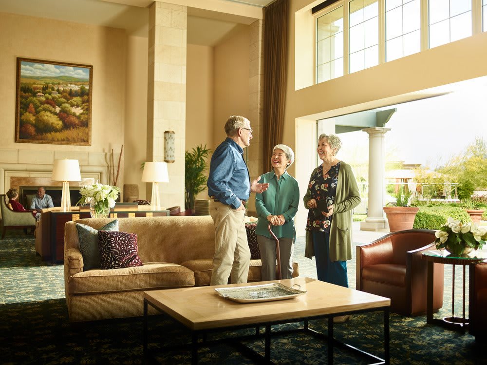 Three people engaged in conversation in a well-lit living room with large windows.