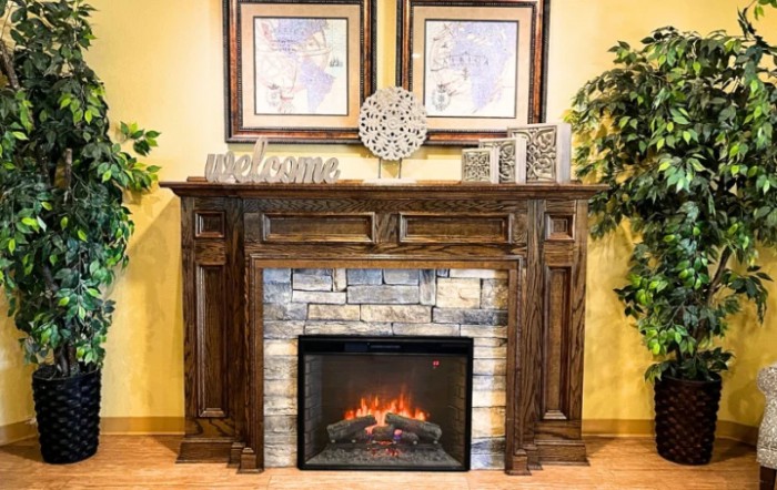 A cozy living room setting featuring an electric fireplace with a wooden mantel, stone surround, and decorative accents including wall art and a clock.