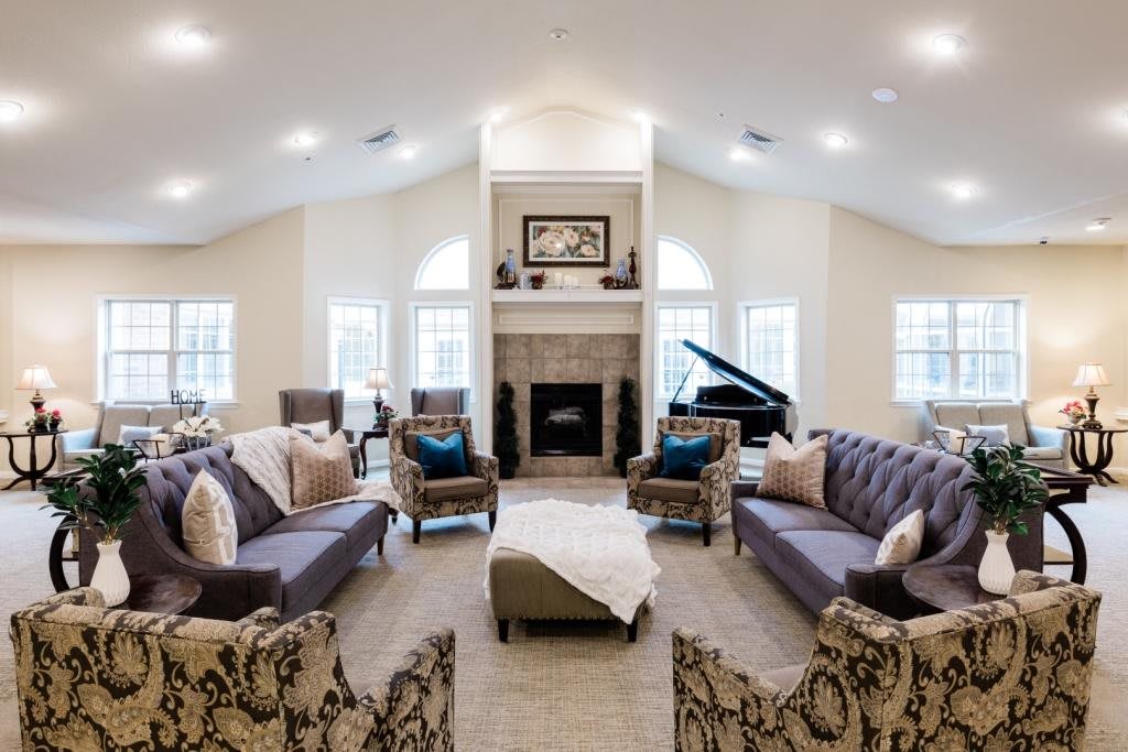 A spacious and elegantly decorated living room featuring a central fireplace, multiple sofas, armchairs, and a grand piano.
