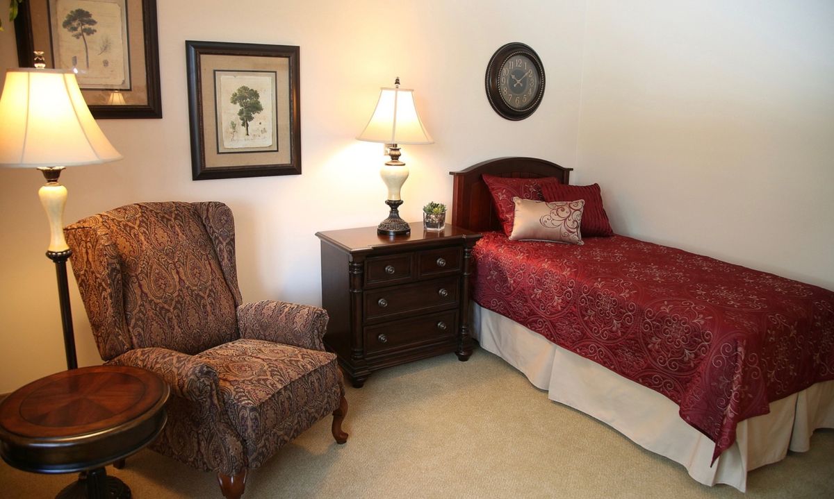 A cozy bedroom with a single bed, nightstand, lamp, and a comfortable chair.
