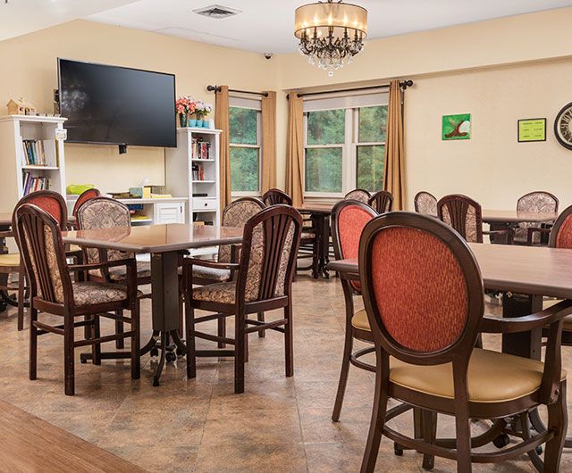 A cozy dining area with several tables and chairs, featuring a television, chandelier, and windows with curtains.