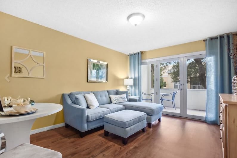 A cozy living room with a blue sectional sofa, wood flooring, and natural light streaming through patio doors.