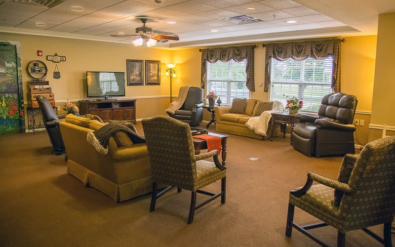 A cozy living room with sofas, chairs, and a TV, featuring large windows and warm lighting.