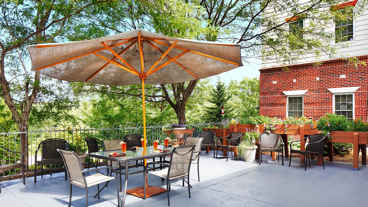 A sunny patio area with outdoor furniture and a large umbrella surrounded by greenery.