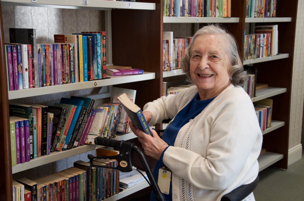 An elderly woman smiling while holding a book in a library.