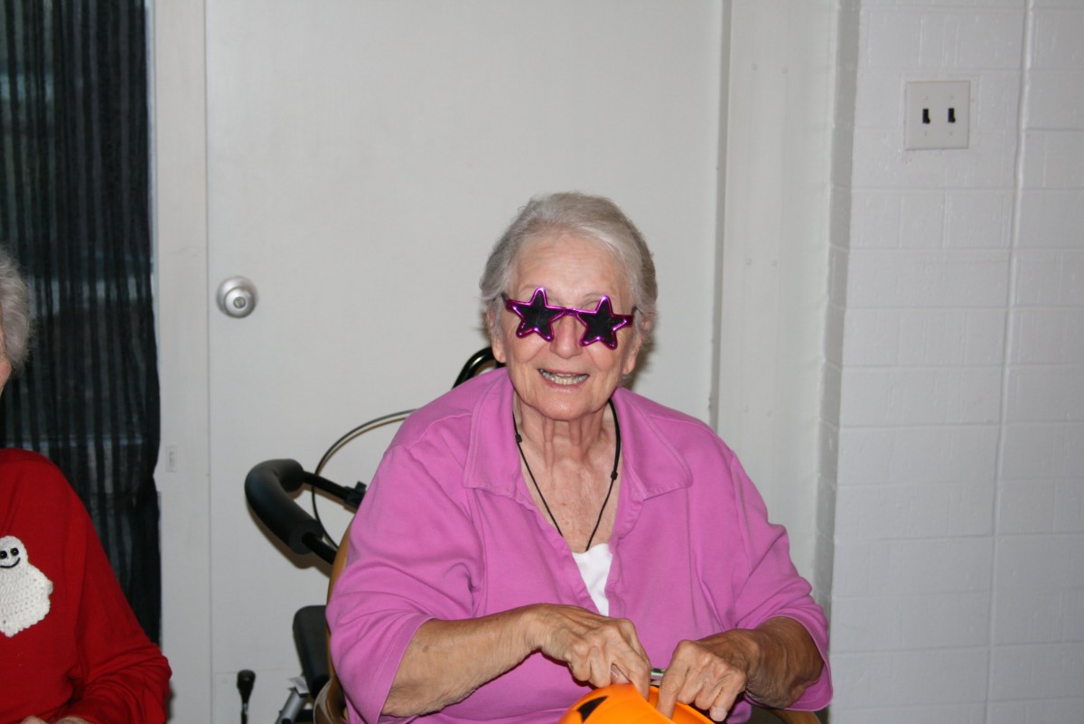 An elderly woman wearing star-shaped sunglasses and a pink shirt, smiling indoors.