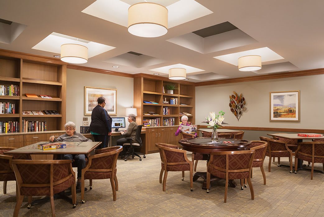 A cozy reading room with elderly individuals engaging in leisure activities amidst bookshelves and comfortable seating.